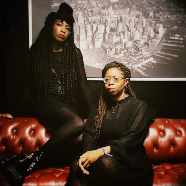Members of Big Joanie sit on a red leather sofa wearing black dresses with a black and white photo of a city behind them.