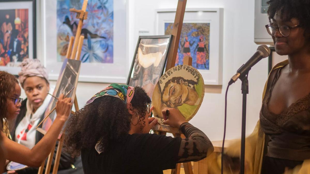 Two women painting at easels whilst another stands at a microphone, with framed art hanging in the background.