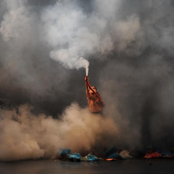 A person stands on a stage shrouded in smoke holding up a flare