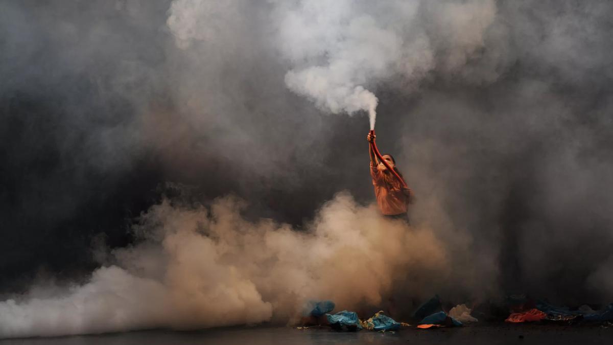 A person stands on a stage shrouded in smoke holding up a flare