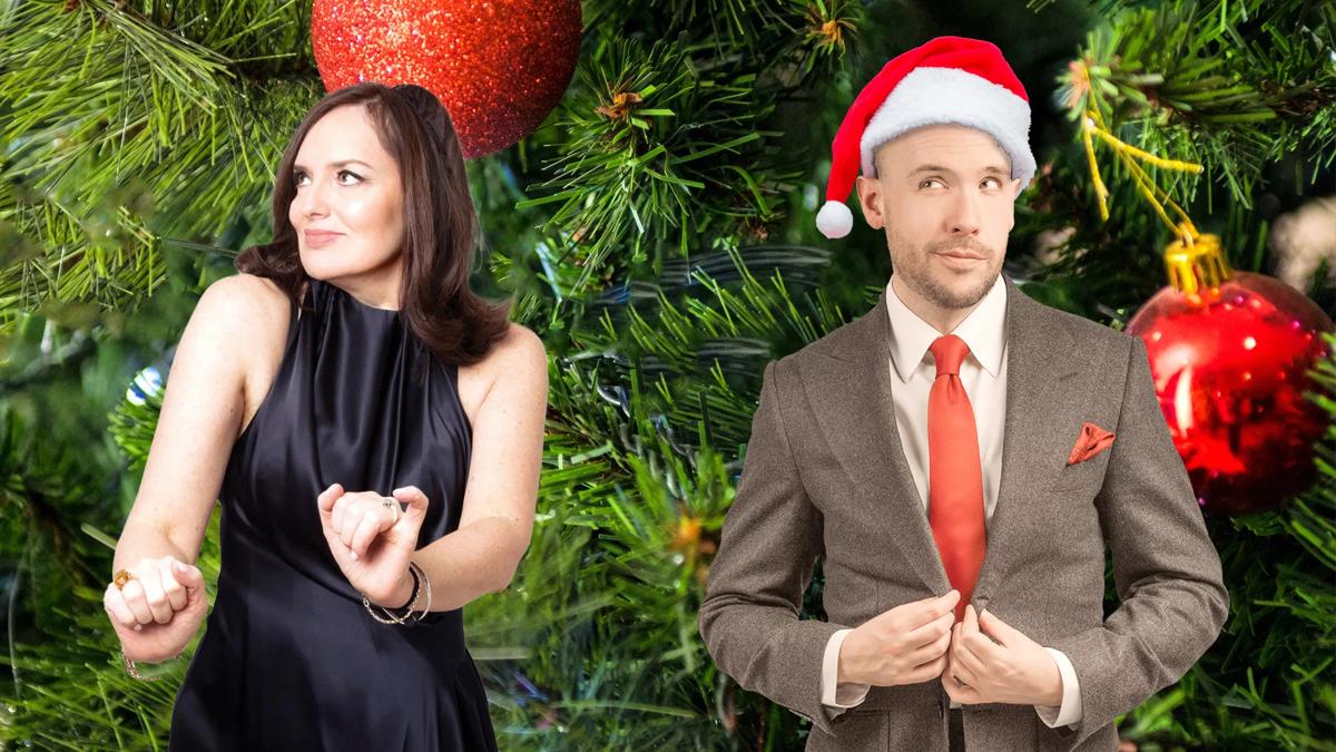 Deborah Frances-White poses mid-shimmy in a black satin dress looking off to the left. Tom Allen stands to her right wearing a brown suit, red tie and a red Santa hat. There is a close up image of branches of a Christmas tree with red shiny baubles in the background. 