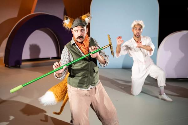 A performer in fox years and tail holds a rake. Behind him another kneels in a white boiler suit.