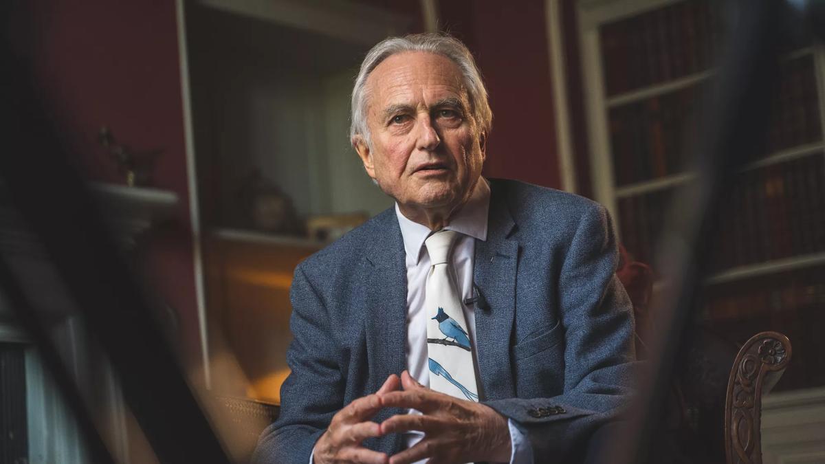 Richard Dawkins wears a blue suit sitting in front of a bookshelf