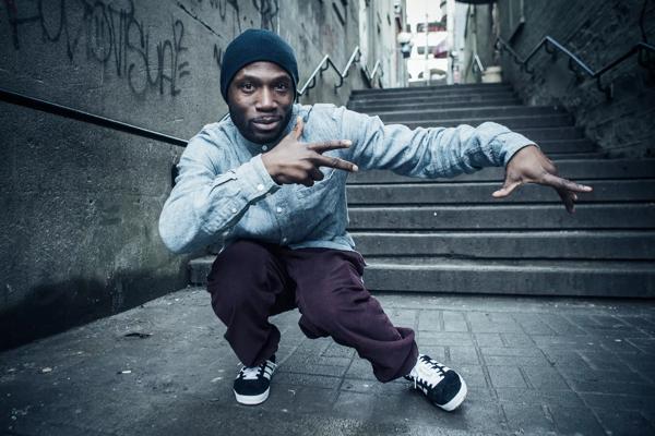 The dancer and choreographer Crazy Smooth. A Black man wearing a collarless denim shirt and a dark blue beanie hat. He is pictured in an urban alleyway. 