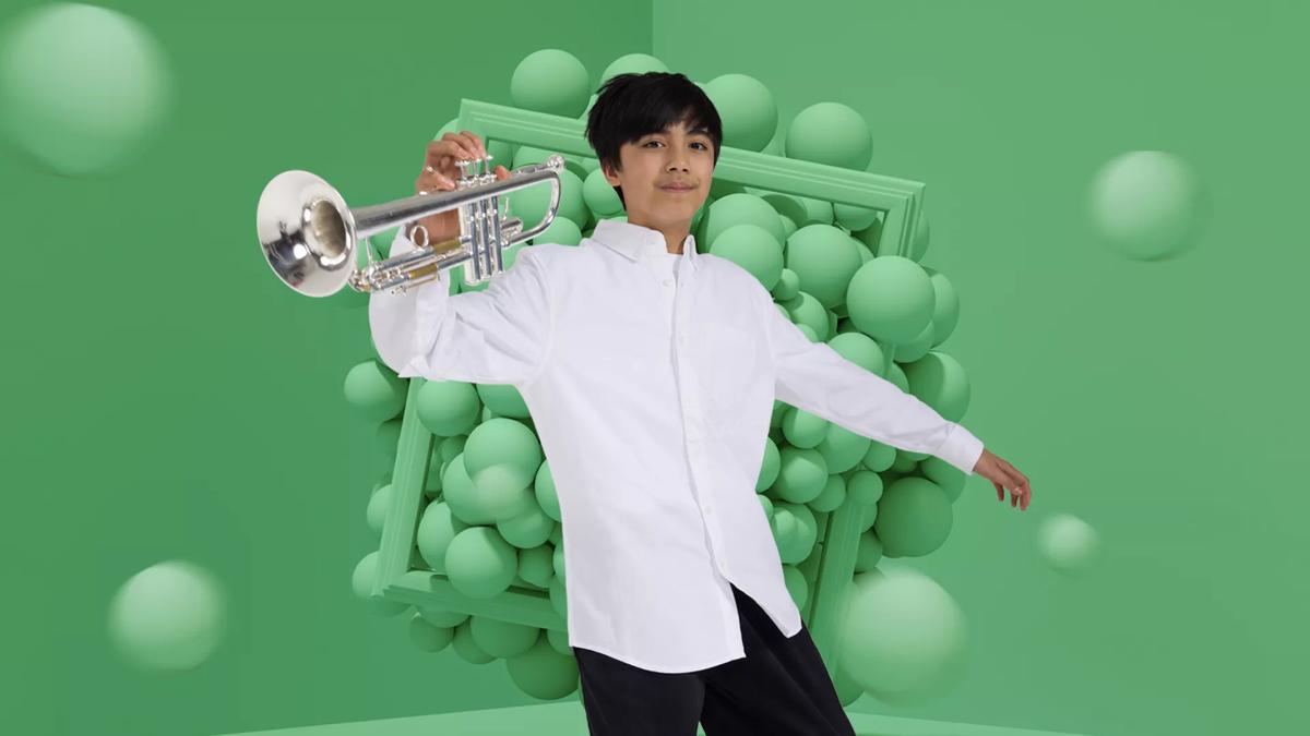 A boy wears a white shirt and black trousers holding a trumpet against a green background with green balls behind him
