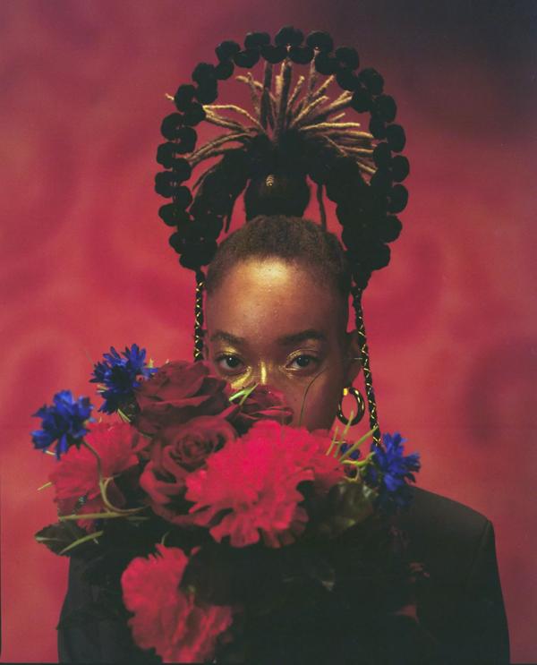 A Black woman with elaborate headdress in front of a crimson red background holds a bouquet of flowers in front of her face