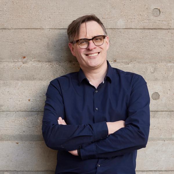 Mark Ball a white man with short dark hair and glasses, stands in front of a concrete wall