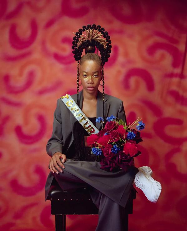 A Black woman seated in front of a patterned red background wears a blue sash and holds a bouquet of flowers
