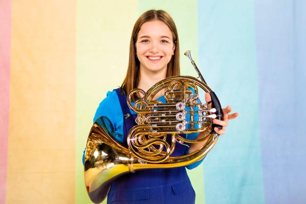 Picture of a girl against a colourful background, holding a brass intstrument. 