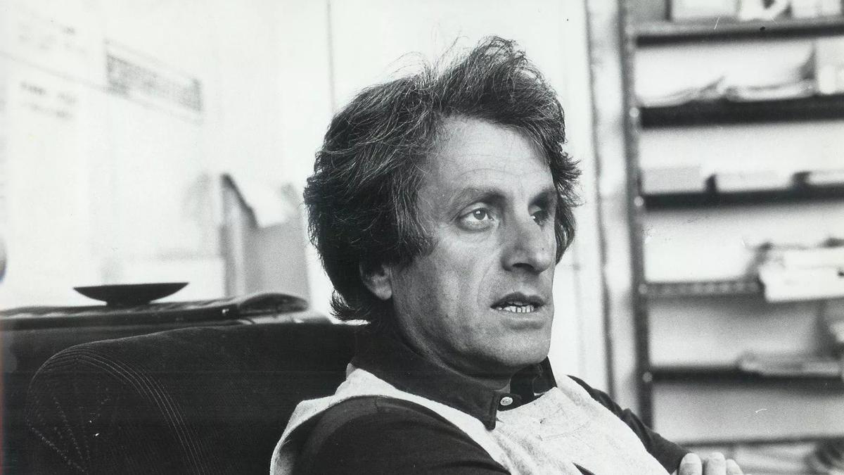 A black and white image of the composer Iannis Xenakis reclining in an armchair in a sparse but untidy study