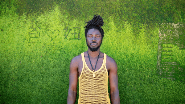Colour portrait of Falle Nioke, who stands against a bright green wall wearing a yellow string vest