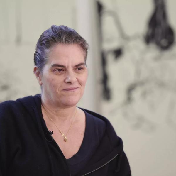 Tracey Emin, a female artist wearing a black fleece fleece jacket over a dark top smiles. Behind her, out of focus, is a piece of artwork