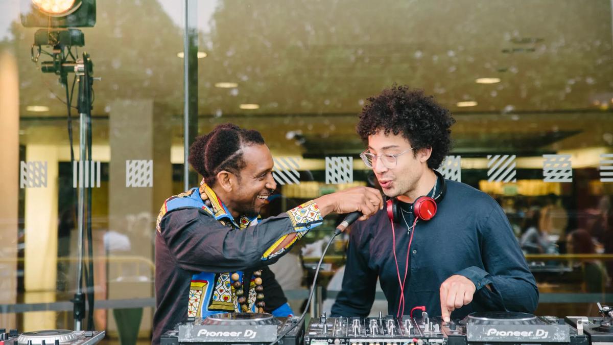 A DJ stands in front of a set of decks while a performer holds a microphone to his mouth.