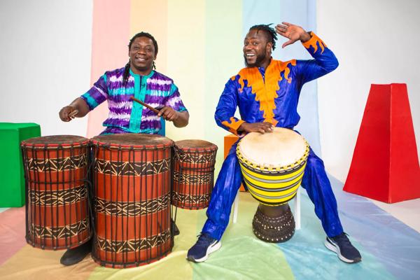Picture of two mean against a colourful background playing African drums. 