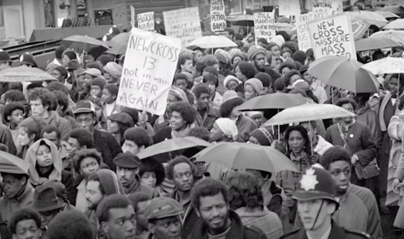 A scene from the 1981 Black People's Day of Action as hundreds of marchers walked from New Cross to Central London following the New Cross Fire; the image features dozens of Black people marching peacefully together some holding umbrellas, others holding placards.