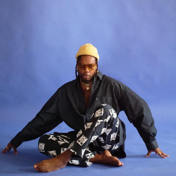 Artist serpentwithfeet sits on the floor cross legged with a yellow beanie hat on in front of a blue back drop.