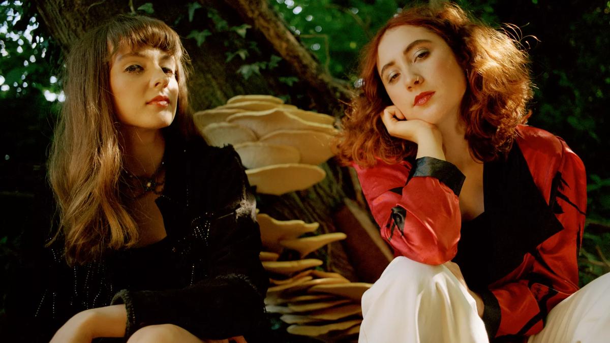 Two women sit in front of a tree with mushrooms growing from it. Both are looking to camera.
