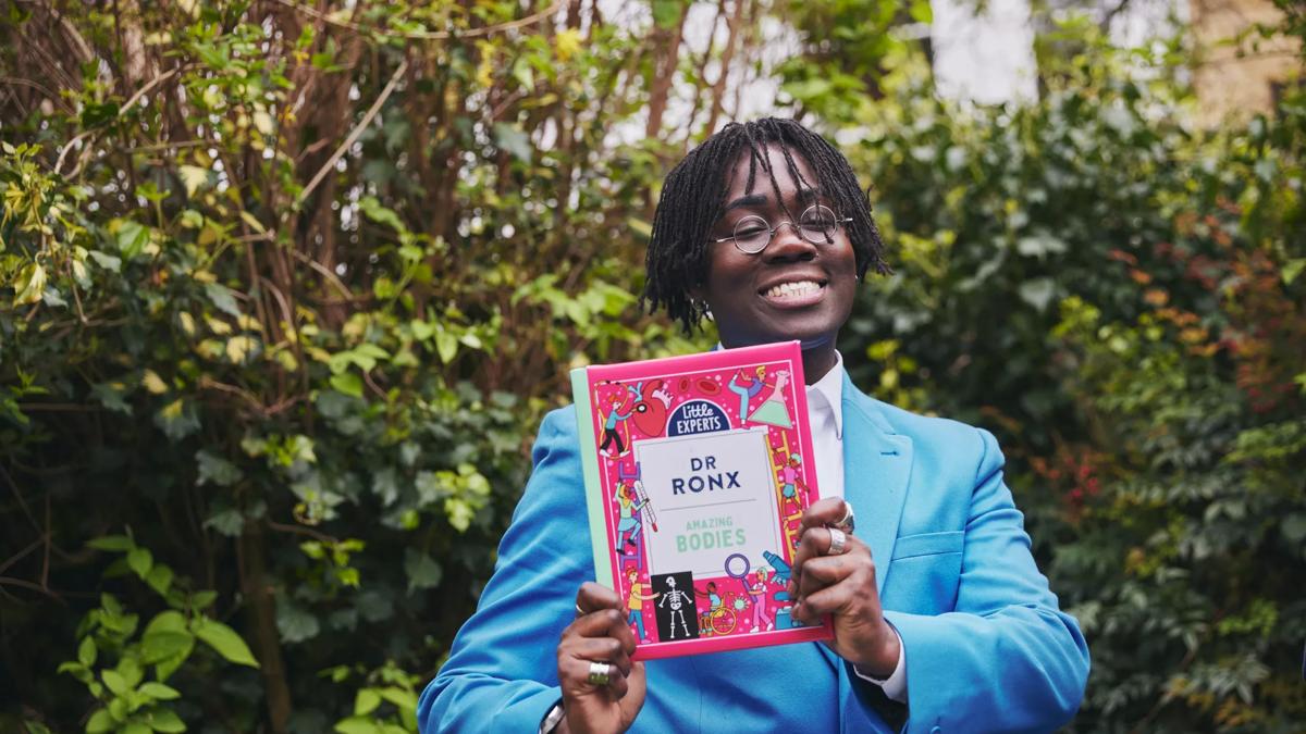 Dr Ronx wears a blue suit in front of a leafy backdrop, holding their book