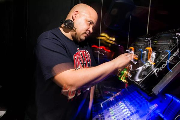 Dj Sugarbear performing a set on the DJ decks wearing a black slogan T-shirt and headphones.