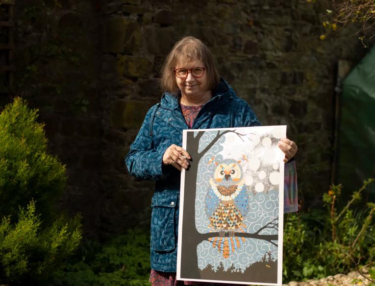 Carol stands outside in a garden holding a loft a piece of artwork depicting an owl sitting on a branch