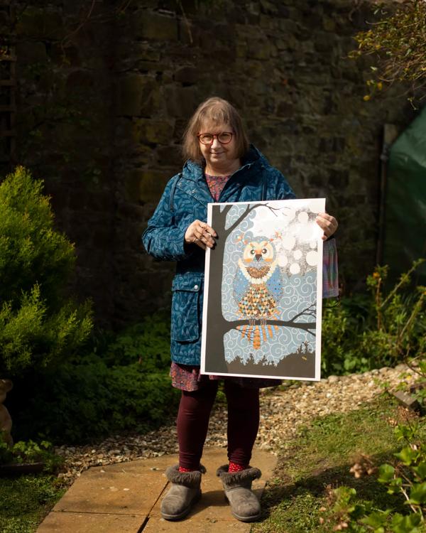Rebecca stands outside in a garden holding a loft a piece of artwork depicting an owl sitting on a branch