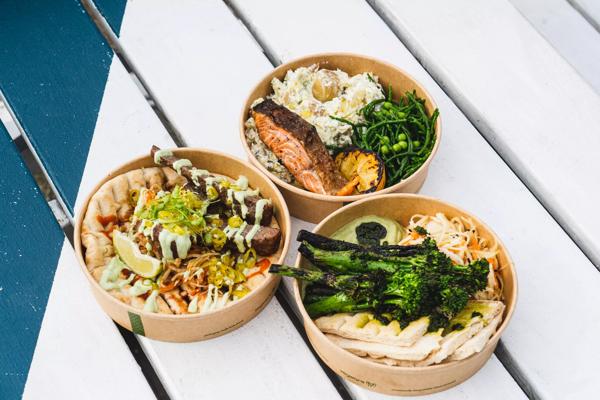 Three paper takeaway bowls. One with potato salad and salmon, one with barbecued broccoli and flatbread, and the third bowl with a flatbread filled with salad