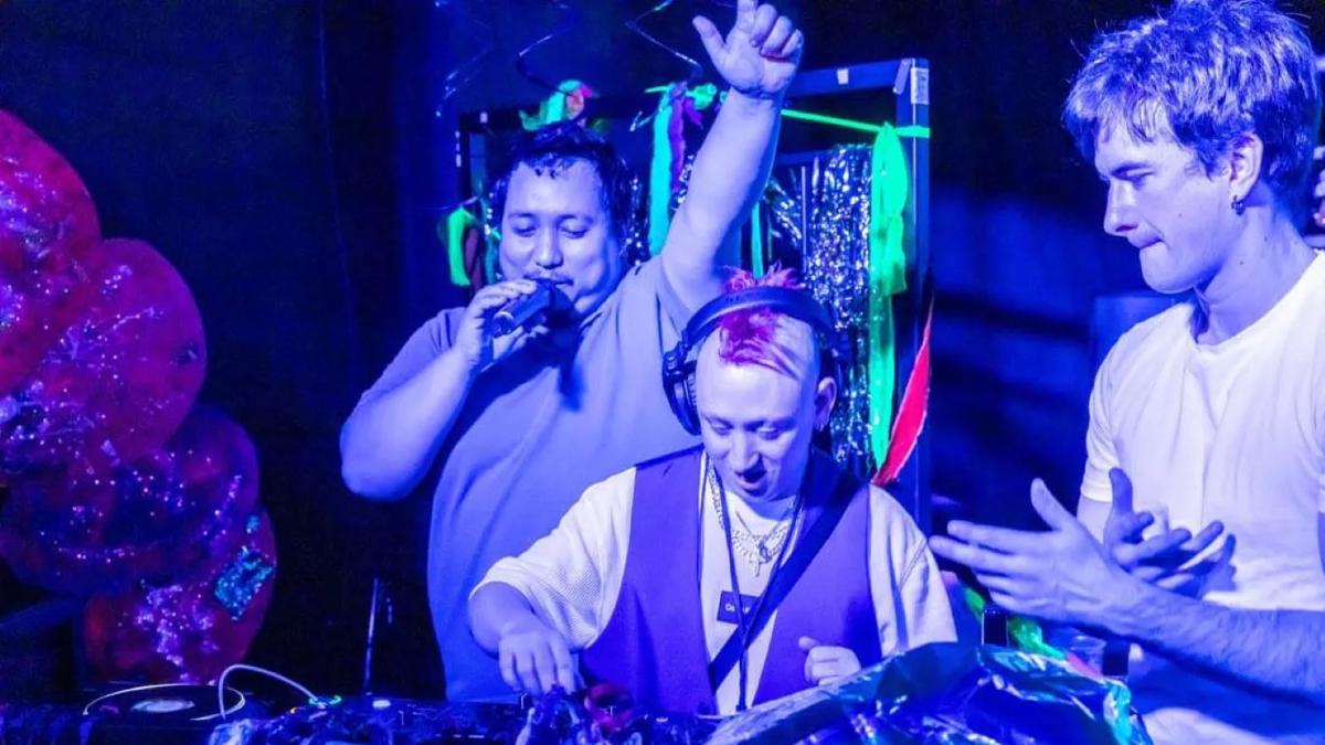 Three people stand around DJ decks lit by blue and purple lighting.