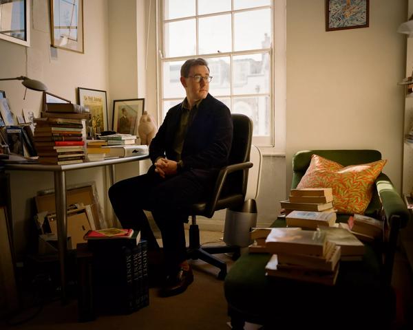 Tom is pictured in his study against a large window. He is surrounded by books and papers. He wears a black coat and trousers and has dark framed glasses. 