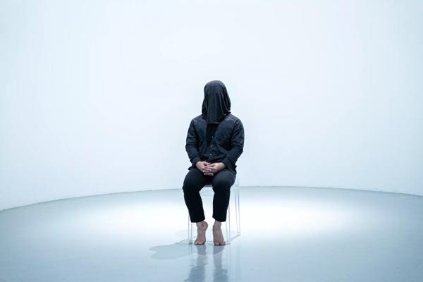Person dressed in black, wearing a black hood over their head, sat down on a transparent chair in a white room. 