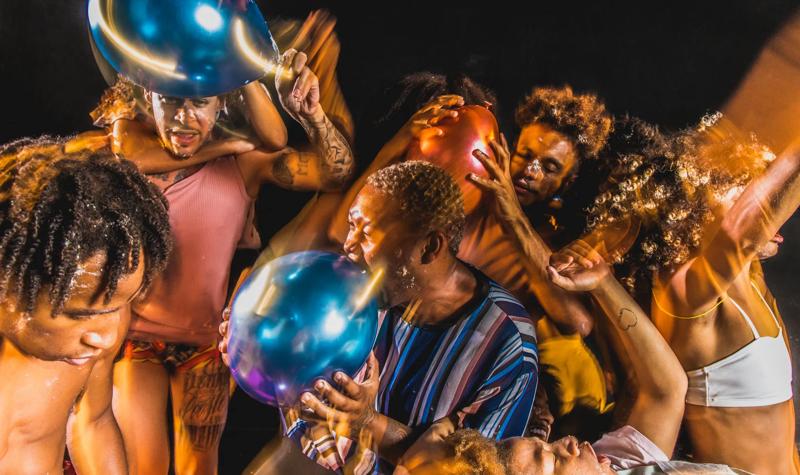 Performers grabbing each other whilst holding and popping balloons 