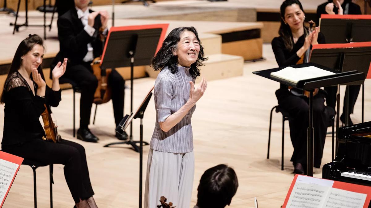 Pianist Mitsuko Uchida standing on stage with the Philharmonia at the Royal Festival Hall, smiling to acknowledge applause, wearing a silver pleated outfit