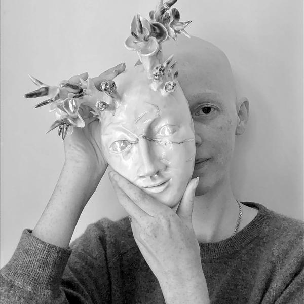 The poet Katie Farris, a young White woman with no hair holds a porcelain mask partly in front of her face