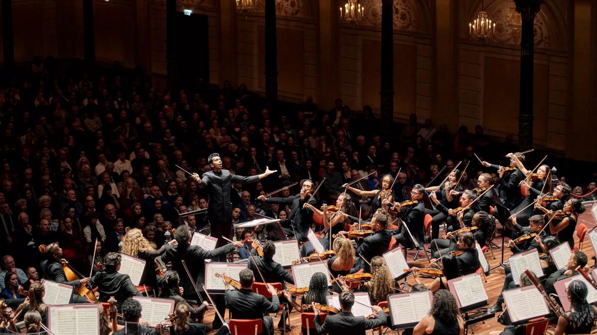 An orchestra on stage with a conductor with his arms open