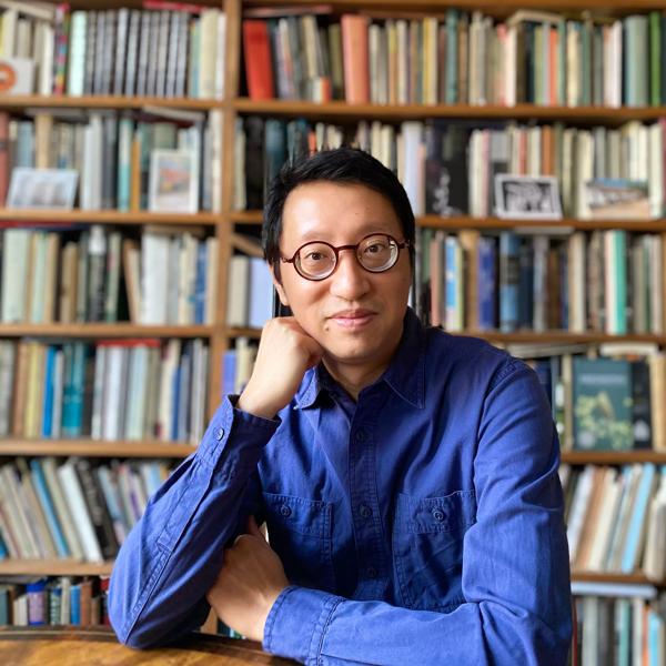 The poet Kit Fan, a middle-aged east Asian man, sits at a table in front of a tall and full bookcase; he wears a blue shirt and round-framed glasses