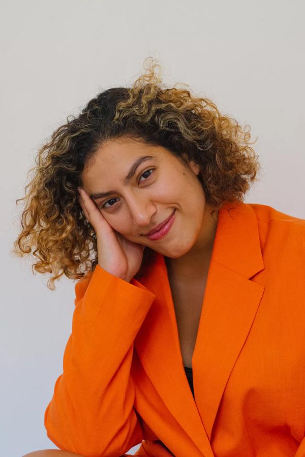 Portrait of tasneim wearing an orange suit jacket, resting their head in their hand and smiling