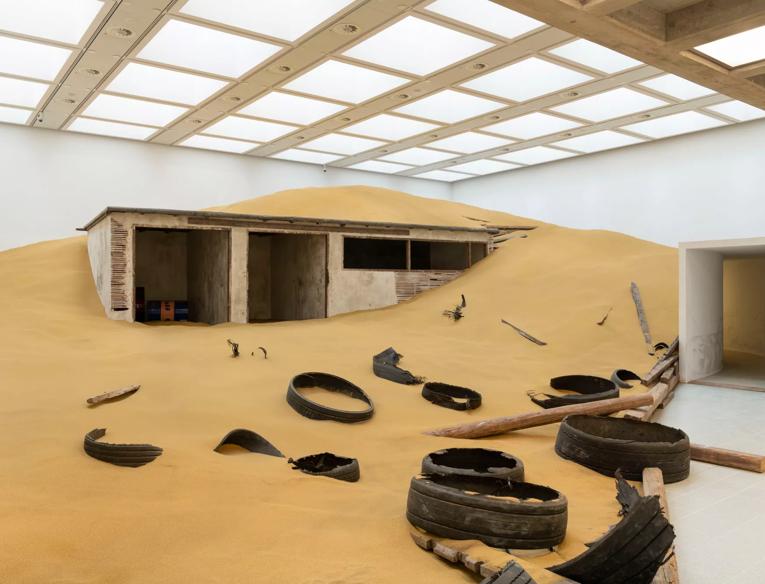 Installation shot of the Mike Nelson exhibition, showing a ruined building half covered in sand with debris strewn about