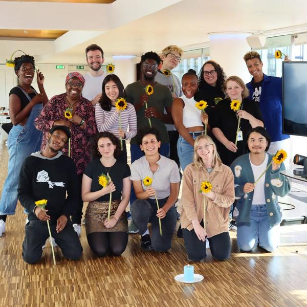 A group of 16 poets pose together (some knelling in front, some standing) next to a large screen with another poet joining the group virtually. They are sitting beding a lit candle, all holding a single sunflower and smiling joyously.