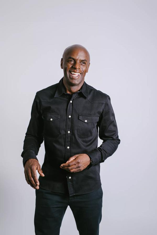 Trevor Nelson wearing black trousers and black button up shirt smiling and standing in front of a white background.