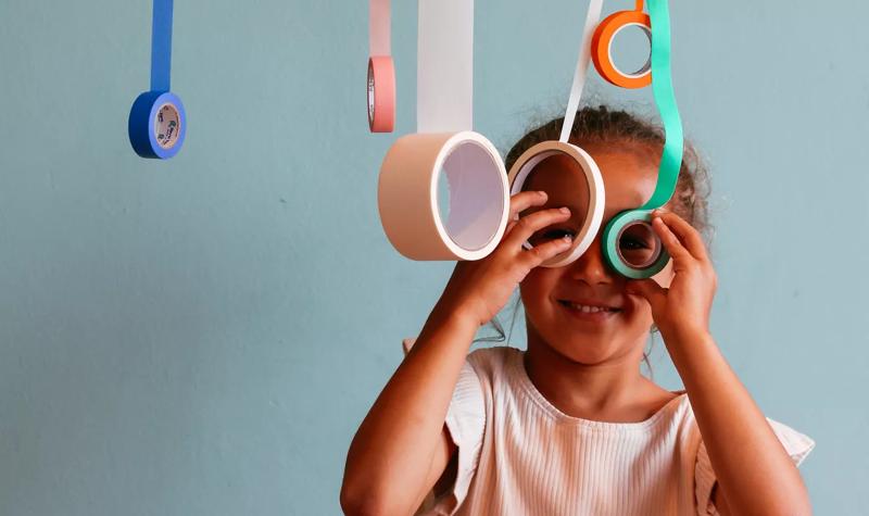 A little girl holds two rolls of tape up to her eyes to look through. Other colourful tapes hang down from the top of  the image. 