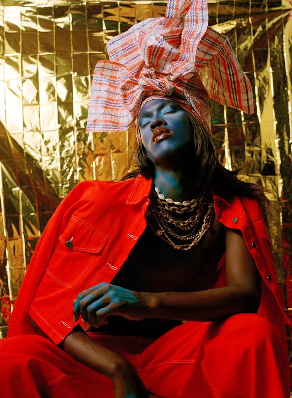A black model in red clothes, blue make-up and styled pink headscarf leans against a gold background