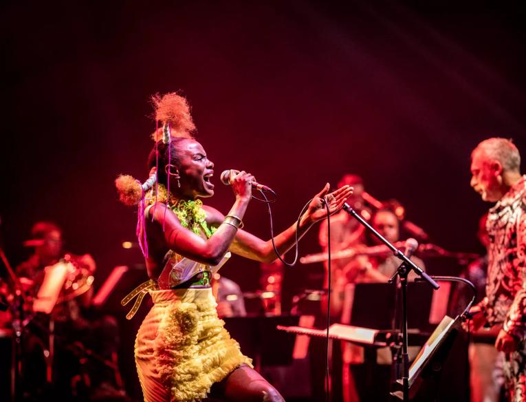Shingai Shoniwa, a black woman in a bright yellow dress, sings on the Royal Festival Hall stage, behind her, out of focus, are members of the Paraorchestra and the orchestra leader Charles Hazlewood, a white man with white hair, in a bright silver suit