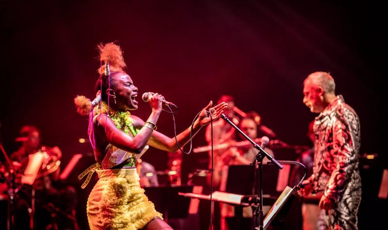 Shingai Shoniwa, a black woman in a bright yellow dress, sings on the Royal Festival Hall stage, behind her, out of focus, are members of the Paraorchestra and the orchestra leader Charles Hazlewood, a white man with white hair, in a bright silver suit
