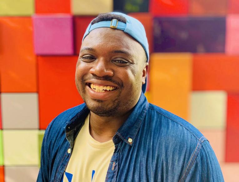 Bryan Washington wears a denim shirt with yellow tshirt underneath and a blue baseball cap. He is photographed against a red, pink and yellow patchwork background. 