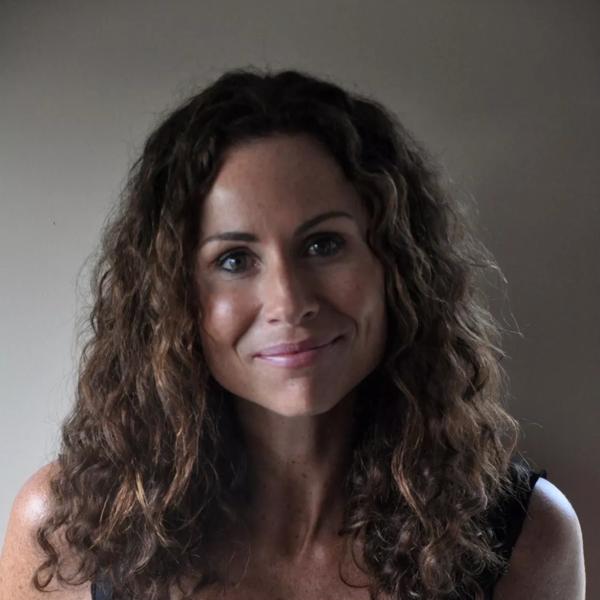 Portrait of author and actress Minnie Driver. She is smiling at the camera and wearing a plain black vest and her brown curls are loose around her shoulders. 