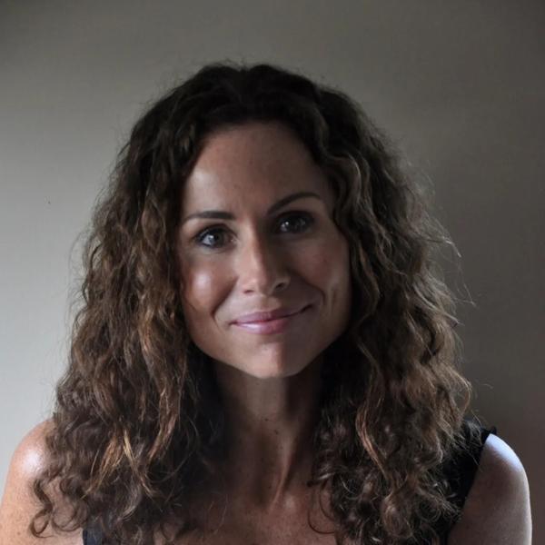 Portrait of author and actress Minnie Driver. She is smiling at the camera and wearing a plain black vest and her brown curls are loose around her shoulders. 