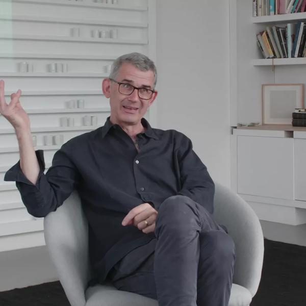 Edmund de Waal, a white man with grey hair, wearing glasses and a dark shirt sits in a chair in a corner of his studio, behind him on one wall are white porcelain objects on shelves in a vitrine, on the other wall are many books on shelves