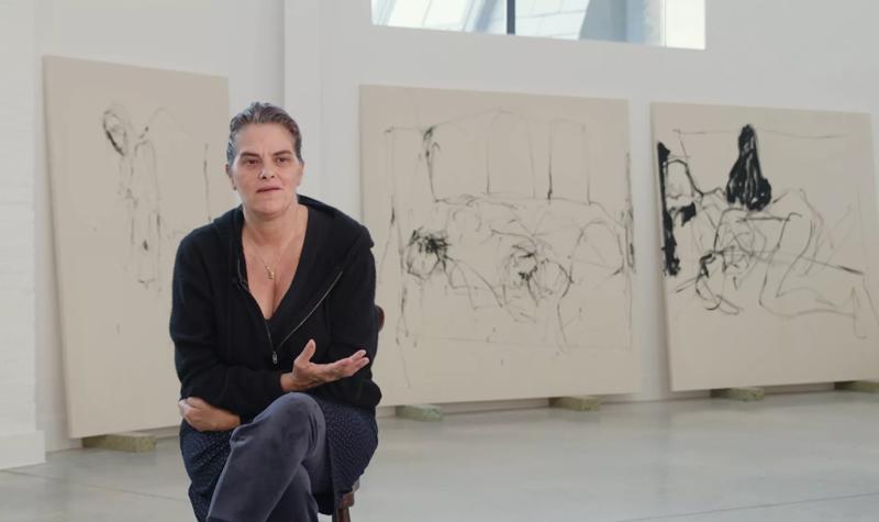 Tracey Emin, a female artist wearing a black fleece fleece jacket over a dark top sits on a chair in her studio. Behind her three large canvases lean against a studio wall