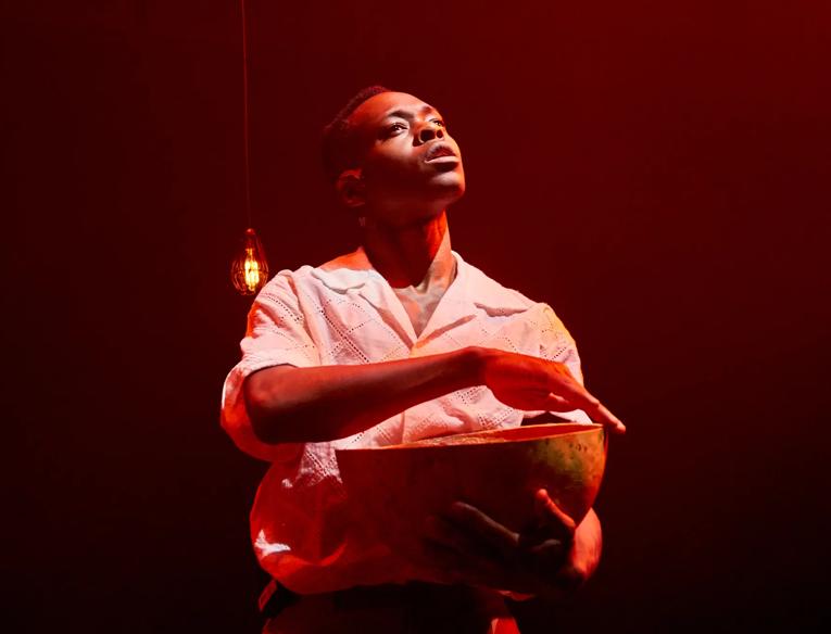 A man holds a wooden bowl while looking off into the distance. He is wearing a white collared short sleeved shirt and is standing in a dimly lit red toned room with a single lightbulb hanging down behind him. 