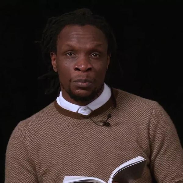 Ishion Hutchinson, a Black man with short dark hair, wearing a brown sweater over a white shirt, reads from their TS Eliot Prize shortlisted collection against a black background