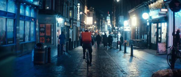 A cyclist with a red backpack cycling past shops with neon signs